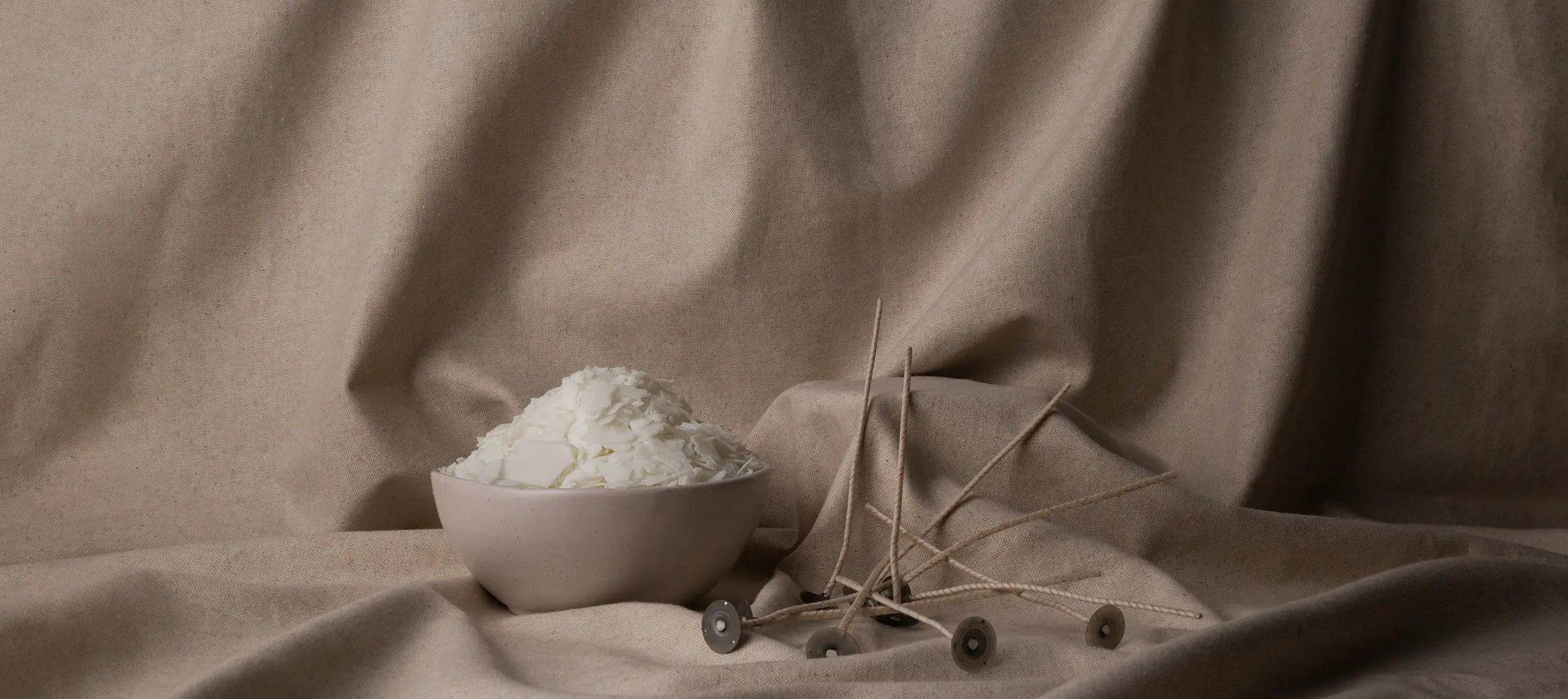 A small bowl of raw soy wax sitting next to a stack of candle wicks on a canvas background